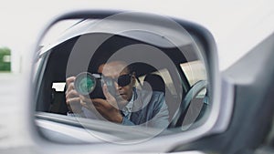 Reflection in side mirror of Paparazzi man sitting inside car and photographing with dslr camera