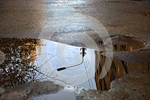 Reflection of the sky, the silhouette of a street lamp and a tree sunlit in a puddle on asphalt