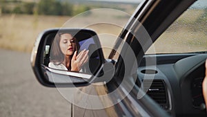 Reflection of woman in car rear view mirror, who applying lip gloss and smiling