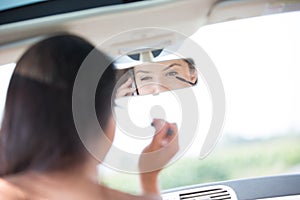 Reflection of woman using cell phone while applying mascara in rearview mirror of car