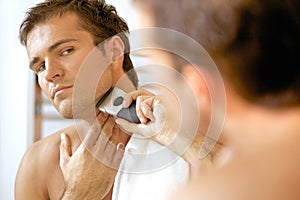 Reflection of young man in mirror shaving with electric shaver