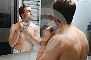 Reflection of young man in mirror shaving with electric shaver