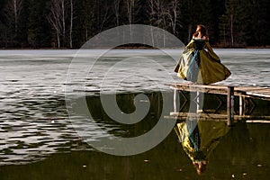 Reflection of a young woman with medieval long dress on the frozen lake in the winter season , forest in the background