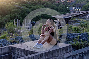 Reflective Dusk A young woman in athletic wear sits thoughtfully on a concrete ledge, with an abandoned building and lush greenery