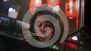 Reflective woman enjoys Chinese meal with chopsticks observing night city street through window