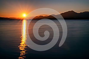 Reflecton of sunrays on water surface. Sun and silhouette of mountains at background