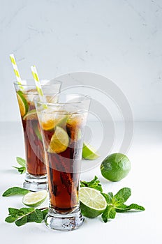 Refreshing alcohol beverage cuba libre with cola, ice, mint, lime, straw in two wet long glasses in modern white kitchen interior,