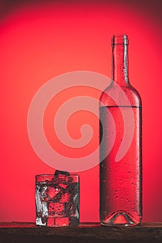 Refreshing cold drink in bottle with drops, and glass with ice on red background. Iced refreshing drinks concept