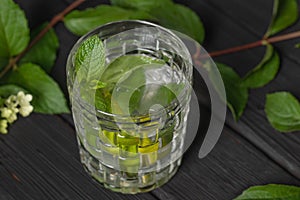 Refreshing cold mojito cocktail with lime and mint in glass on burnt black wood background decorated with summer flowers.