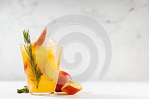 Refreshing juicy peach cocktail with ice, rosemary, sugar rim, fruit slices in misted glass, ingredients on white wood table.