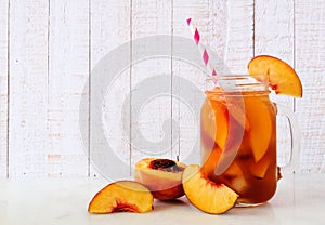 Refreshing peach iced tea in a mason jar glass with cut fruit against white wood