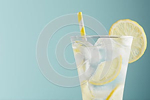 Refreshing summer tonic water with lemon, ice cubes, yellow straw in light soft pastel blue color background, closeup, half, top.
