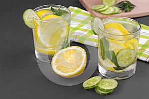 Water with lemon and cucumber and mint in a glass cup next to half a fresh lemon and cucumber slices on a black background