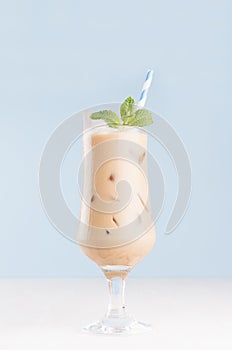 Refreshment baileys creamy cocktail in misted elegant wineglass with ice cubes, green mint, straw in light blue interior on white.