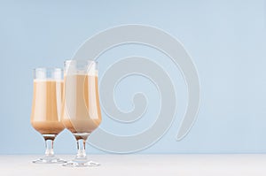Refreshment cold coffee drink in two goblets with cream and foam in modern light pastel blue interior on white wood table.