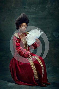 Regal Beauty. Elegant medieval royal person, young african woman, queen in stylish dress with fan sitting against