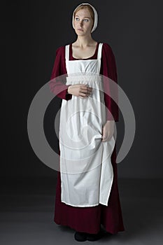 A Regency maid servant wearing a red linen dress with an apron and cap against a studio backdrop