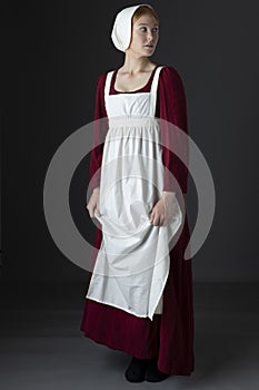 A Regency maid servant wearing a red linen dress with an apron and cap against a studio backdrop