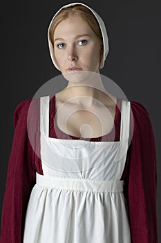 A Regency maid servant wearing a red linen dress with an apron and cap against a studio backdrop