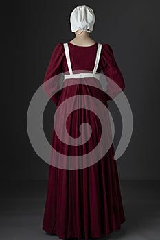 A Regency maid servant wearing a red linen dress with an apron and cap against a studio backdrop