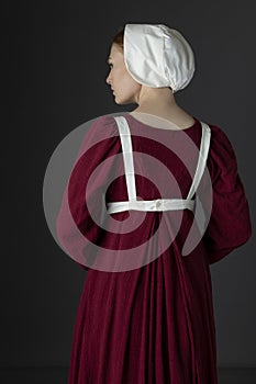 A Regency maid servant wearing a red linen dress with an apron and cap against a studio backdrop