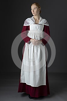 A Regency maid servant wearing a red linen dress with an apron and cap against a studio backdrop