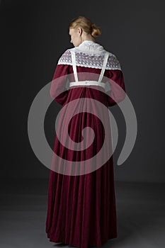 A Regency maid servant wearing a red linen dress with an apron and a lace modesty shawl against a studio backdrop