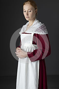 A Regency maid servant wearing a red linen dress with an apron and a lace modesty shawl against a studio backdrop
