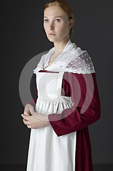 A Regency maid servant wearing a red linen dress with an apron and a lace modesty shawl against a studio backdrop