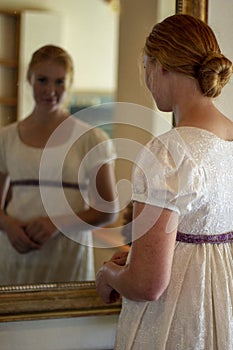 Regency woman in cream dress looks in a gold framed mirror