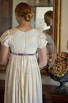 Regency woman in cream dress stands before a mirror