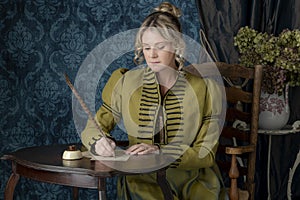 A Regency woman wearing a green linen spencer and sitting at a small table writing with a quill