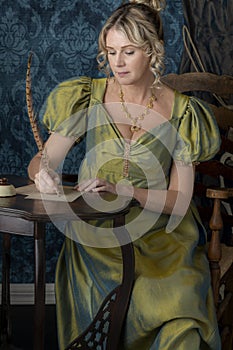 A Regency woman wearing a green shot silk dress and sitting at a table writing a letter with a quill