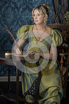 A Regency woman wearing a green shot silk dress and sitting at a table writing a letter with a quill