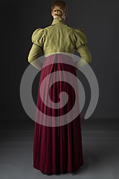 A Regency woman wearing a  red linen dress with a green spencer with black braid  against a studio backdrop