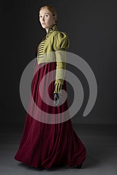 A Regency woman wearing a  red linen dress with a green spencer with black braid  against a studio backdrop