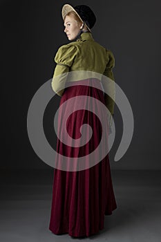 A Regency woman wearing a red linen dress with a green spencer with black braid plus gloves and a bonnet against a studio backdrop