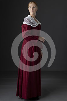 A Regency woman wearing a  red linen dress with  a lace modesty shawl against a studio backdrop
