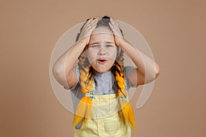 Regretting young child with yellow kanekalon pigtails, touching head with hands and with worried face in yellow jumpsuit