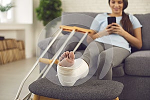 Woman with broken leg in plaster cast sitting on couch at home with mobile in hands