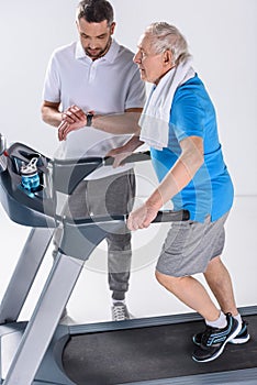 rehabilitation therapist checking time while assisting senior man exercising on treadmill