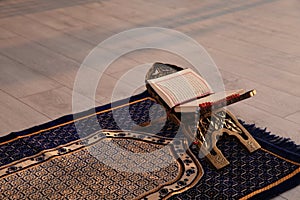 Rehal with open Quran and Muslim prayer beads on rug indoors.