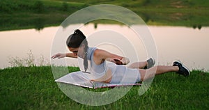 A rejuvenated woman in the background of a lake in a park lying on a gymnastic mat on her stomach performs a body lift