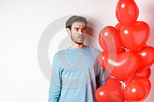 Relationship. Young man looking confused at heart balloon, puzzled on valetines day, standing over white background