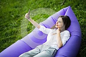 Relax on fresh air. Pretty young woman lying on inflatable sofa lamzac use mobile phone while resting on grass in park on the sun