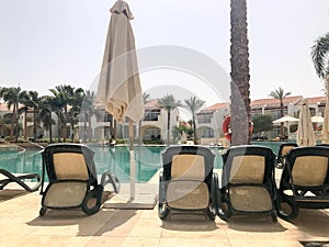 Relax pool with clear blue water with sunbeds for sunbathing with sun umbrellas against the backdrop of green palms and white buil