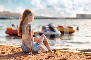 Relax at resort. Young caucasian smiling tanned woman with tattoos poses sitting at the sand beach. Ocean and jet skis