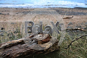 Relax Sign sitting on driftwood with beach and sea behind