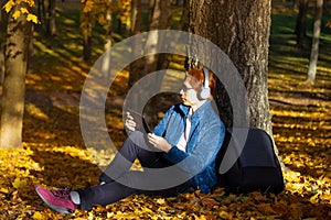 Asian young man  lean the tree and looking Tablet as a listening to music in autumn season with a sunny light in the