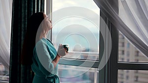 relax time at home at weekend, woman is looking at window, enjoying city view and drinking hot tea
